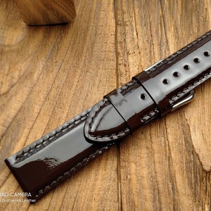 Pu&ograve; includere: Un cinturino da orologio in pelle marrone scuro con finitura lucida. Il cinturino ha un bordo cucito e una fibbia argentata.