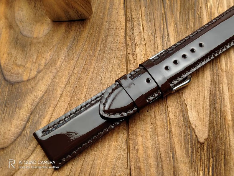 Pu&ograve; includere: Un cinturino da orologio in pelle marrone scuro con finitura lucida. Il cinturino ha un bordo cucito e una fibbia argentata.