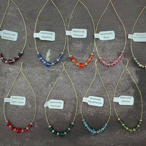 May include: A collection of Swarovski crystal necklaces in various colors, including amethyst, sapphire, fireopal, rose, light siam, emerald, aquamarine, olivine, and vitral medium. Each necklace features a delicate gold-colored wire and a small tag with the crystal name.