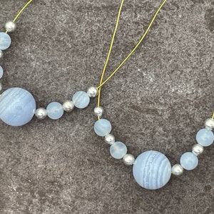 May include: Two strands of beads, each with a large, light blue, striped gemstone bead, smaller light blue beads, and silver-coloured beads. The beads are strung on a thin, gold-coloured wire against a grey background.
