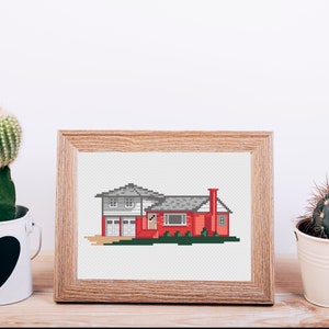 May include: A framed cross-stitch of a red house with a gray roof and a small green lawn. The house has a garage door and a chimney. The frame is made of wood and has a light brown finish.
