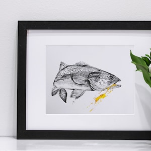 May include: A black and white drawing of a fish with gold paint splatters on a white background. The drawing is framed in a black frame and is displayed on a white shelf.