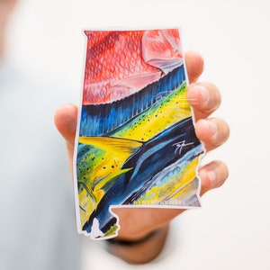 May include: A sticker featuring a colorful illustration of a mahi-mahi fish in the shape of the state of Alabama. The fish is depicted in shades of red, yellow, blue, and green. The sticker includes the text "The Foxishly".
