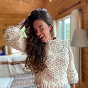 May include: A woman wearing a white chunky knit sweater with a high neckline and a relaxed fit. She is smiling and has long, curly brown hair. She is wearing blue jeans and has a gold bracelet on her wrist.
