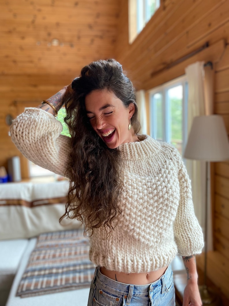 May include: A woman wearing a white chunky knit sweater with a high neckline and a relaxed fit. She is smiling and has long, curly brown hair. She is wearing blue jeans and has a gold bracelet on her wrist.