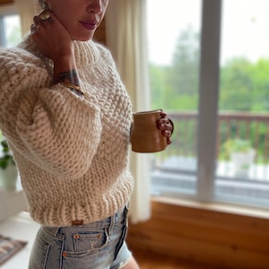 May include: A close-up of a woman wearing a cream-colored chunky knit sweater with a high neck and balloon sleeves. She is holding a brown ceramic mug.