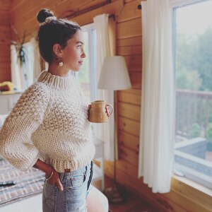 May include: A woman wearing a cream-colored knitted sweater and denim shorts stands in a cabin, holding a brown ceramic mug. She is looking out a window with white curtains.