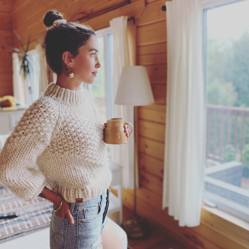 May include: A woman wearing a cream-colored knitted sweater and denim shorts stands in a cabin, holding a brown ceramic mug. She is looking out a window with white curtains.