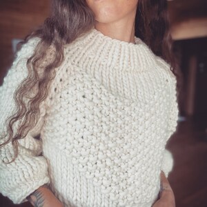 May include: A white knitted sweater with a chunky knit texture. The sweater has a high neckline and a relaxed fit.