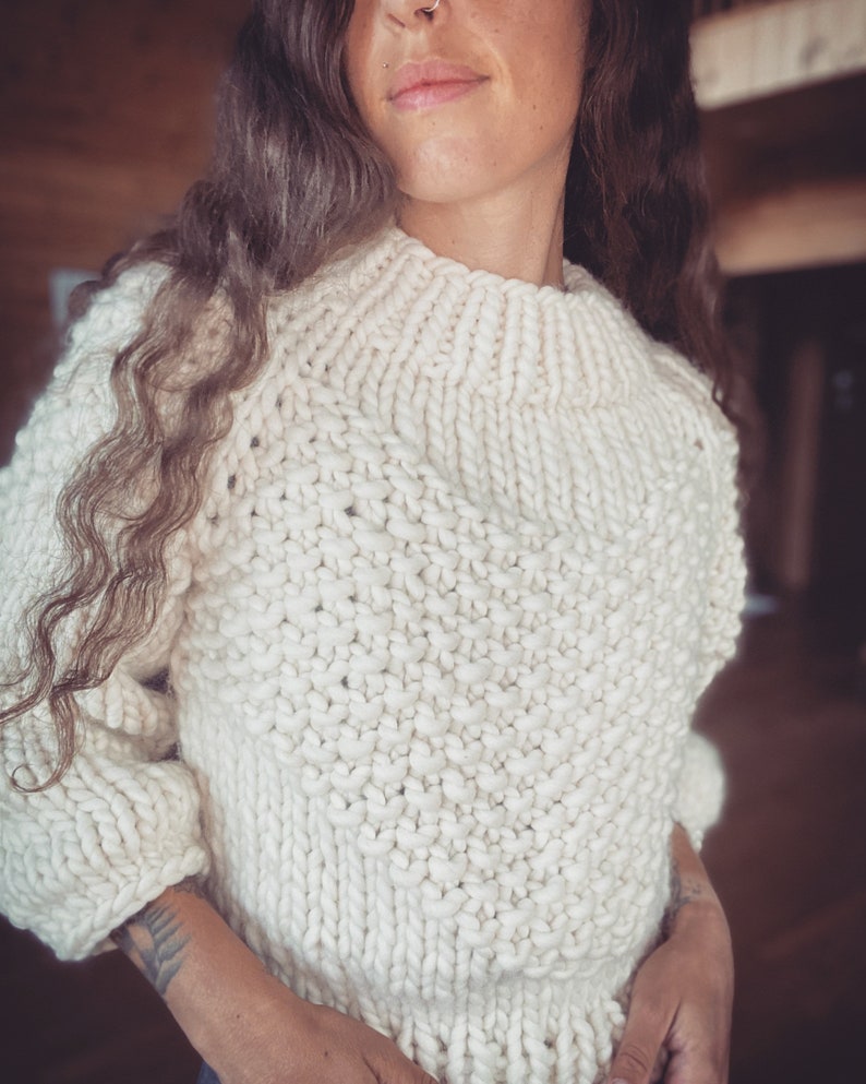 May include: A white knitted sweater with a chunky knit texture. The sweater has a high neckline and a relaxed fit.