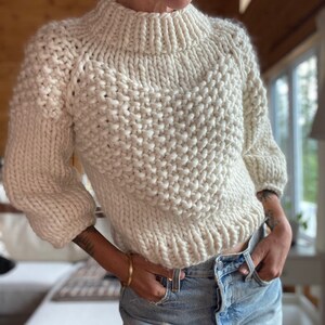 May include: A white chunky knit sweater with a high neck and a slightly cropped length. The sweater is made with a thick yarn and has a textured stitch pattern.