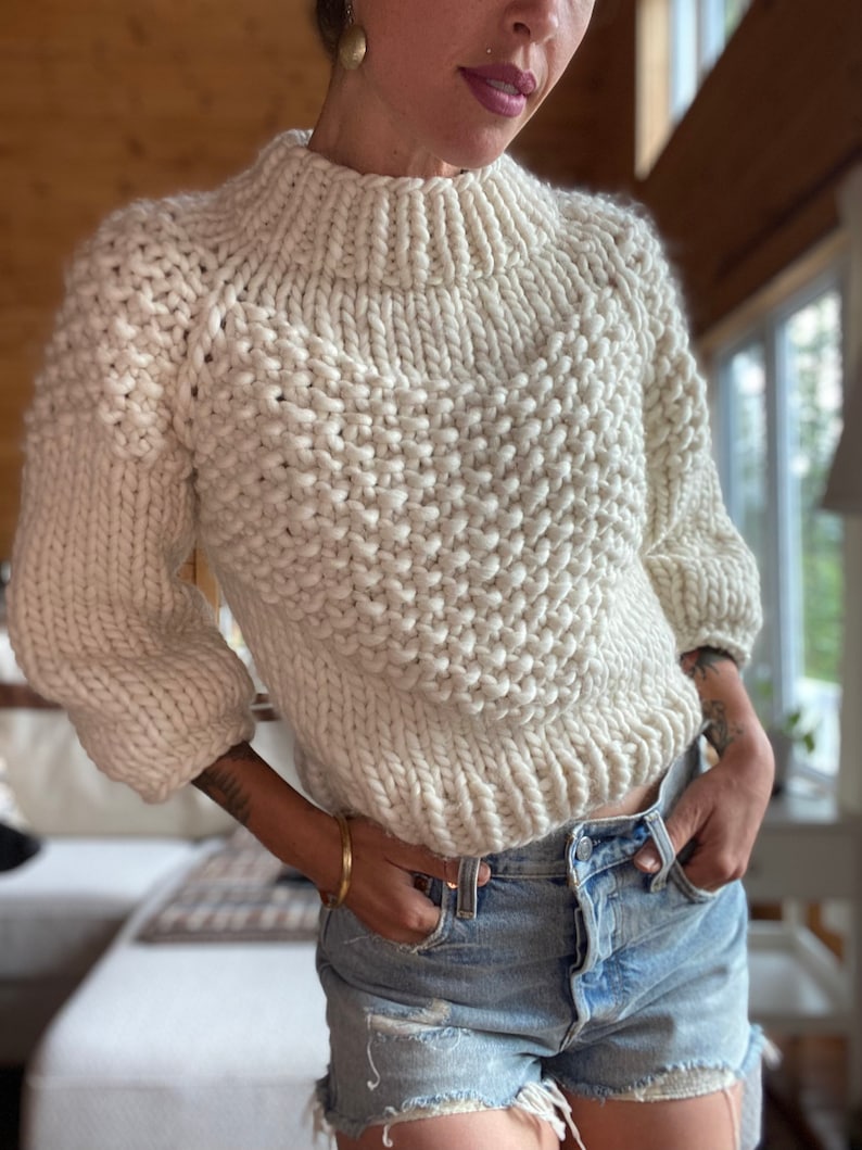 May include: A white chunky knit sweater with a high neck and a slightly cropped length. The sweater is made with a thick yarn and has a textured stitch pattern.
