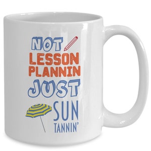 May include: White ceramic coffee mug with the text "NOT LESSON PLANIN' JUST SUN TANNIN'" in blue, red, and orange. A yellow and blue striped beach umbrella is below the text.