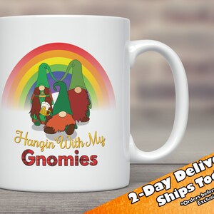 May include: White ceramic mug featuring a rainbow and three gnomes holding a beer. The text "Hangin' With My Gnomies" is printed on the mug in a red and black tartan font.