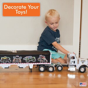 May include: A young boy is playing with a large white toy semi-truck and trailer. The trailer is black and has four monster truck decals on the side. The text "Decorate Your Toys!" is in a white box with an orange background.