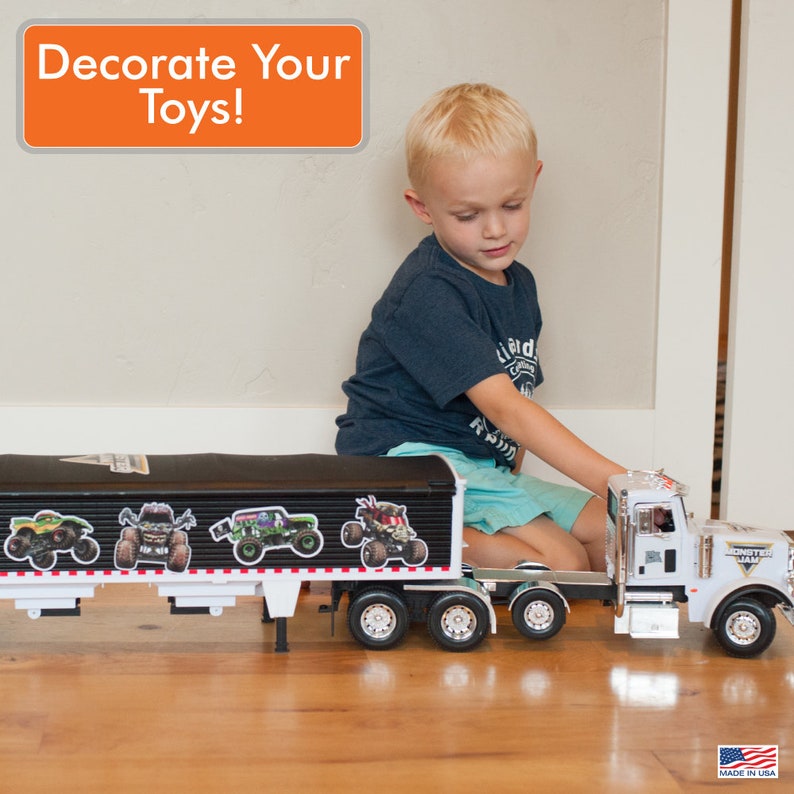 May include: A young boy is playing with a large white toy semi-truck and trailer. The trailer is black and has four monster truck decals on the side. The text "Decorate Your Toys!" is in a white box with an orange background.