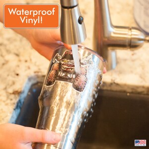 May include: A stainless steel tumbler with a colourful sticker of a cartoon monster on it. Water is being poured over the sticker to show that it is waterproof vinyl.