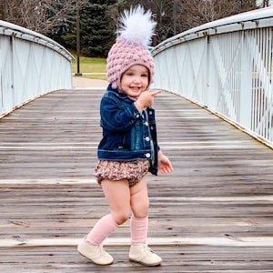 May include: A young child wearing a pink crocheted hat with a white pom-pom, a denim jacket, a floral print skirt, and pink knee-high socks. The child is standing on a wooden bridge and pointing to the right.