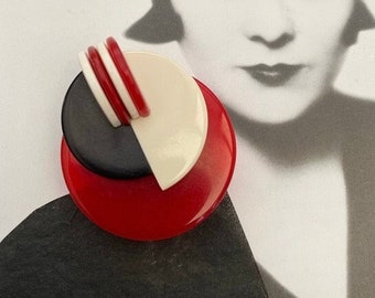 May include: A red, white, and black geometric brooch with a layered design. The brooch is made of plastic and has a circular shape.