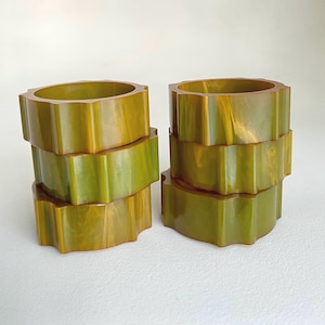 Peut inclure: Deux piles de ronds de serviette en résine vert olive et jaune. Chaque anneau a un design unique à bord festonné. Les anneaux sont empilés par groupes de trois, mettant en valeur leurs variations de forme et de couleur. Le fond est blanc.