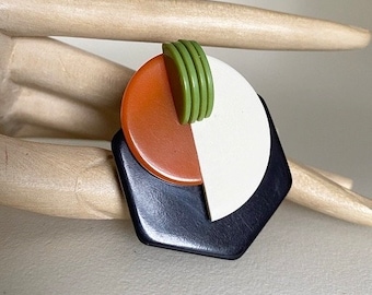 May include: A geometric brooch with a white, orange, and green design. The brooch is made of plastic and has a black backing.