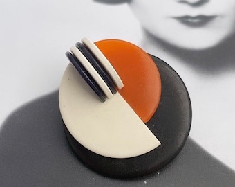 May include: A round, black and white geometric brooch with a bright orange accent. The brooch is made of resin and has a modern art deco style.