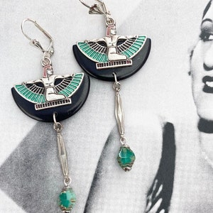 May include: Silver-tone earrings with a black, semi-circular design depicting a seated figure with outstretched wings, highlighted with teal and red. Each earring has a dangling silver bar with a teal bead.