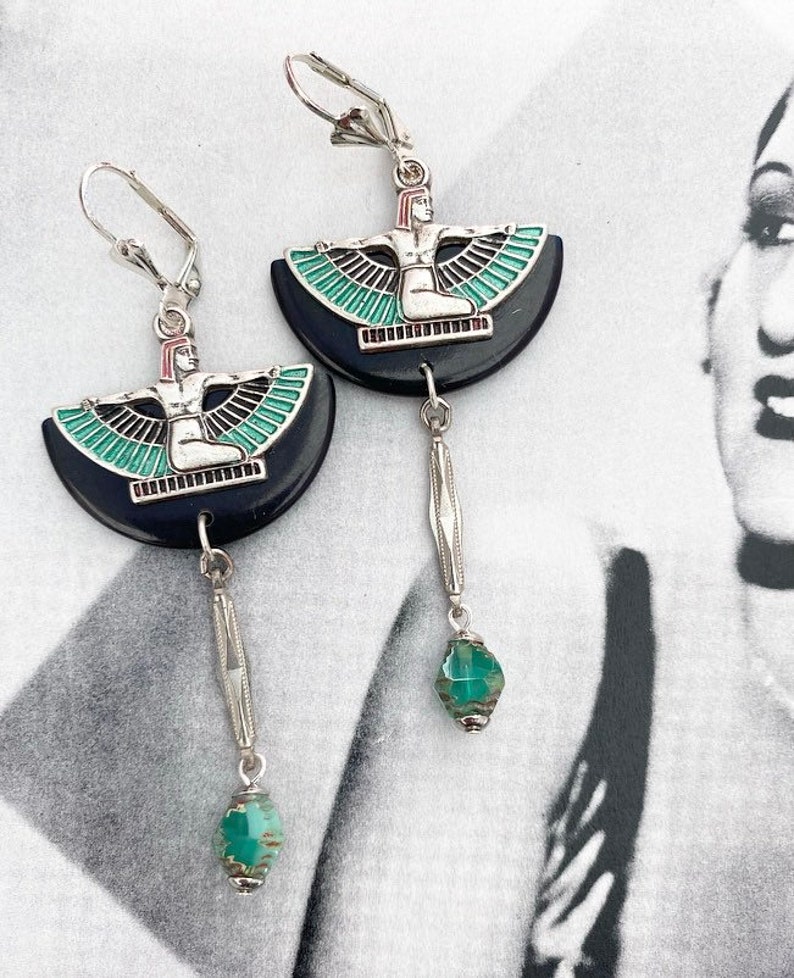 May include: Silver-tone earrings with a black, semi-circular design depicting a seated figure with outstretched wings, highlighted with teal and red. Each earring has a dangling silver bar with a teal bead.