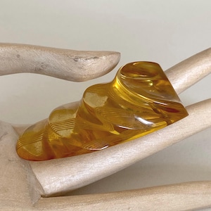 Può includere: Un anello traslucido color ambra con un design unico a forma di onda. La superficie dell'anello presenta sottili linee parallele, creando un effetto strutturato. L'anello è esposto su una mano di legno.