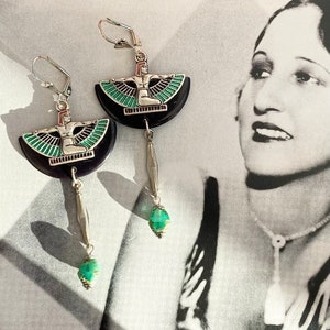 May include: Silver earrings with an Egyptian design. Each earring has a black, semi-circular base with a silver figure and green accents. A dangling silver element and a green gemstone complete the design. The earrings are set against a vintage black and white photograph.
