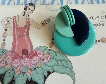 May include: A geometric brooch composed of layered circular and semi-circular resin discs in shades of mint green, teal, and dark navy blue. The design features a large teal base disc topped with a smaller mint green disc and a dark navy blue semi-circle.