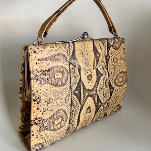 May include: A structured handbag featuring a textured snakeskin pattern in shades of tan, beige, and dark brown. The bag has a single top handle, a silver-toned metal frame, and a spherical clasp closure at the top center of the rectangular body.