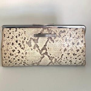 Op de afbeelding: Een rechthoekige clutch met een slangenpatroon in crème en bruin. De clutch heeft een zilverkleurig metalen frame en sluiting. Het slangenpatroon is gedetailleerd en bedekt het gehele oppervlak.