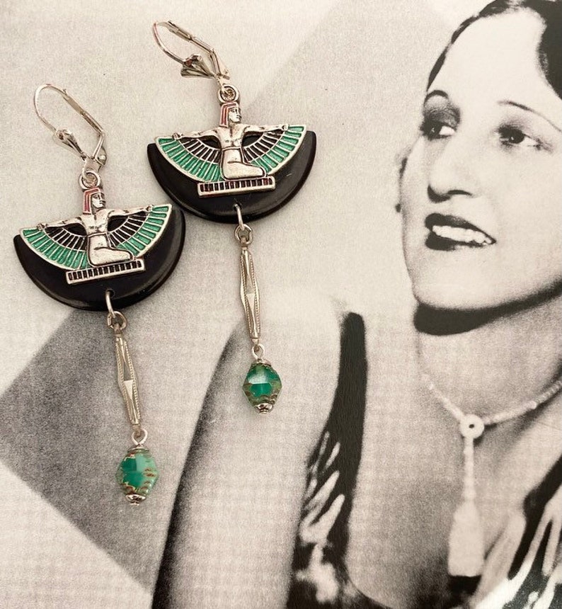 May include: Silver leverback earrings. Each earring features a black, semi-circular design with a silver and enamel figure. A dangling silver bar and a faceted, teal bead complete the design. The earrings are approximately 6 cm long.