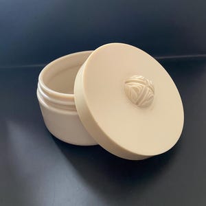 May include: A white round container with a lid. The lid has a raised, decorative design in the centre.