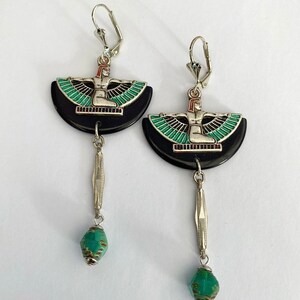 May include: A pair of silver leverback earrings. Each earring has a black, semi-circular pendant with a silver figure and green and black wings. A silver bar and a turquoise bead dangle from each pendant.