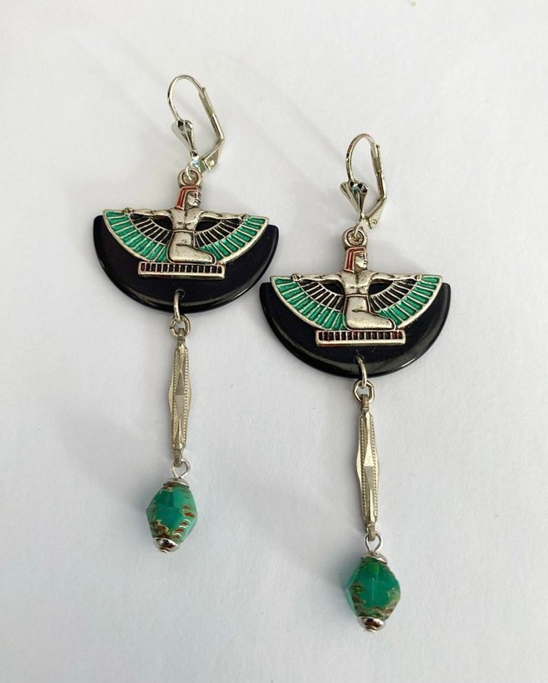 May include: A pair of silver leverback earrings. Each earring has a black, semi-circular pendant with a silver figure and green and black wings. A silver bar and a turquoise bead dangle from each pendant.