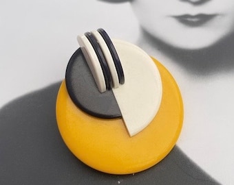 May include: A yellow, black, and white geometric brooch with a circular design. The brooch is made of plastic and has a modern art deco style.