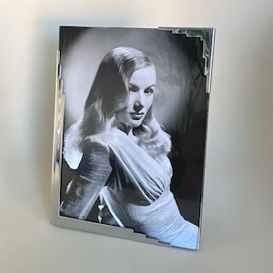 Könnte beinhalten: Schwarz-Weiß-Fotografie einer Frau in einem silbernen Rahmen. Der Rahmen ist rechteckig mit dekorativen Ecken. Das Foto zeigt eine Frau mit welligem Haar und einem Kleid.