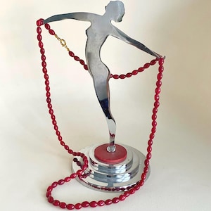 May include: A silver metal sculpture of a woman holding a red beaded necklace. The necklace is draped across the sculpture's outstretched arms. The sculpture is mounted on a chrome base with a red accent.