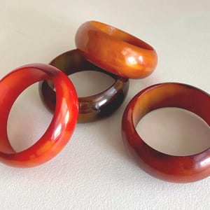 Może przedstawiać: Cztery gładkie, zaokrąglone bransoletki typu bangle w różnych odcieniach bursztynu, pomarańczu i głębokiego czerwonawego brązu. Każda bransoletka ma polerowane, półprzezroczyste wykończenie z subtelnymi zmianami koloru w materiale, ułożone w grupę.