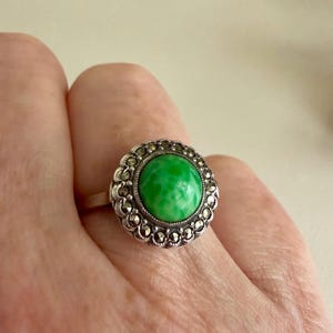 May include: A silver ring featuring a round, bright green cabochon stone at the centre. The stone is surrounded by a decorative border of small, dark, faceted marcasite stones set in a scalloped metal frame with a polished silver finish.