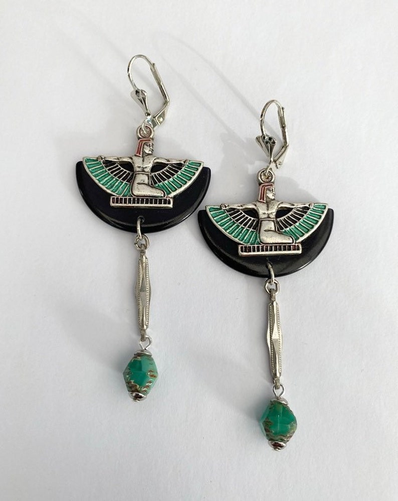 May include: A pair of silver earrings with a leverback clasp. Each earring has a black, semi-circular base with a silver figure and green and black wing design. A silver bar and a green bead hang from each earring.
