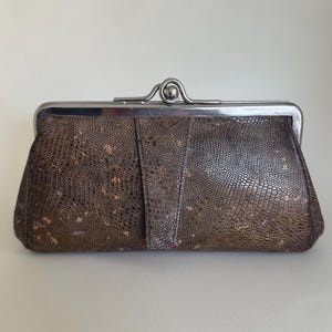 May include: A rectangular clutch bag featuring a dark brown, textured surface with a repeating small-scale pattern and subtle metallic gold-toned flecks. The bag is secured with a silver-toned metal frame and a central ball-clasp closure.