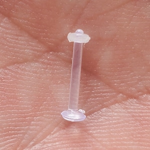 May include: A clear plastic body piercing with a small, round top and bottom. The piercing is about 1 inch long.
