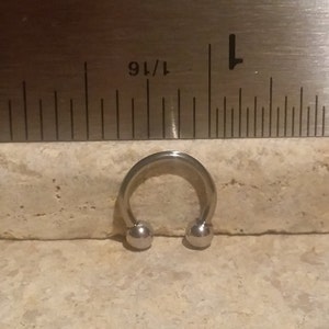 May include: A silver circular barbell with two ball ends, measuring 1/16 inch in diameter, is shown next to a ruler.