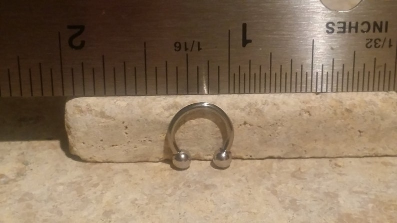 May include: A silver circular barbell with two ball ends, measuring 1/16 inch in diameter, is shown next to a ruler.