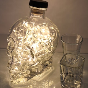 May include: A clear glass bottle shaped like a skull with fairy lights inside. The bottle is sitting on a table with two shot glasses, one with the text "Jack Daniel's Whiskey".