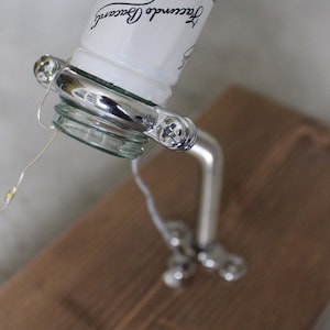 May include: A clear glass bottle of white rum with a silver liquor pour spout. The bottle is angled, with the label visible. The spout is attached to a silver stand with decorative accents. The stand is on a brown wooden surface.