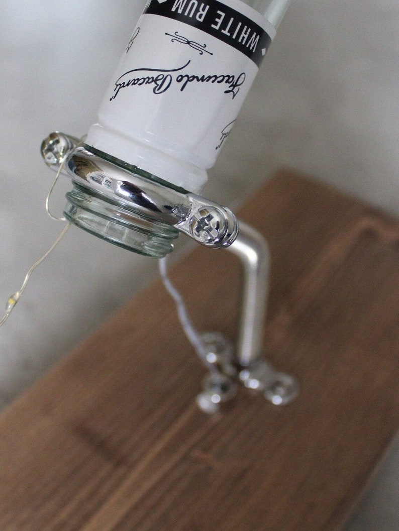 May include: A clear glass bottle of white rum with a silver liquor pour spout. The bottle is angled, with the label visible. The spout is attached to a silver stand with decorative accents. The stand is on a brown wooden surface.